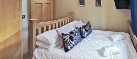 Una cama bien hecha con sábanas blancas y almohadas estampadas, un cabecero de madera, una puerta de madera, una ventana pequeña y un televisor de pantalla plana montado en la pared.