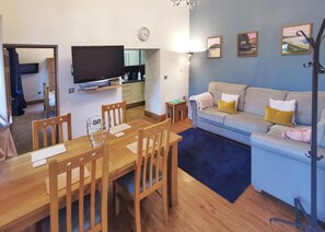 A living room with a sofa, a dining table with chairs, a TV, a clock, and a small side table. - Carmarthenshire Cottage (Clunderwen, Nr Narberth)