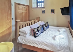 Ein ordentlich bezogenes Bett mit weißen Leinen und gemusterten Kissen, ein hölzerner Kopfteil, eine Holztür, ein kleines Fenster und ein an der Wand befestigter Flachbildfernseher.