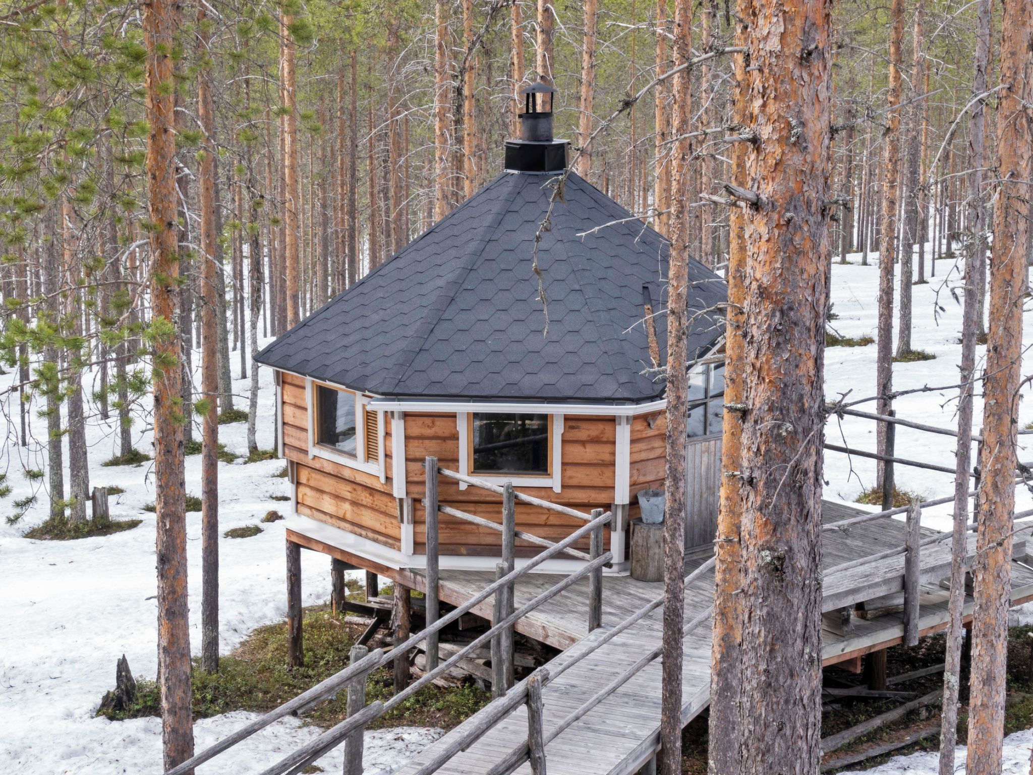 Una cabaña de madera con techo hexagonal, rodeada por un bosque con nieve en el suelo.