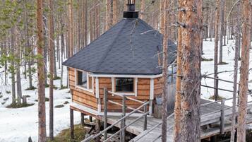 Una cabaña de madera con techo hexagonal, rodeada por un bosque con nieve en el suelo.