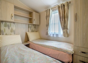 A bedroom with two beds, a wardrobe, and a window with curtains. - 12ft-wide luxury caravan with one double and one twin. (Charmouth)