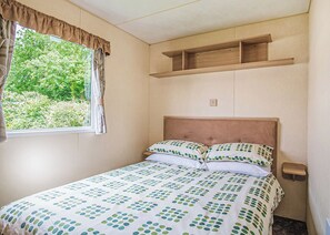 A single bed with a patterned bedspread, a window with curtains, and a wooden shelf above the bed. - Spacious and comfortable 12ft-wide caravan with one double and two twin. (Cross Inn, Nr New Quay)