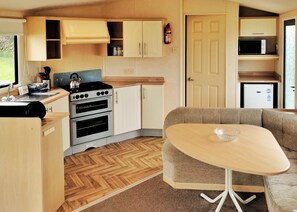 A compact kitchen with a stove, oven, and microwave, a dining area with a table and sofa, and a door leading to another room. - Spacious and comfortable 12ft-wide caravan with one double and two twin. (Cross Inn, Nr New Quay)