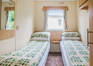 A compact bedroom with two single beds, a window with curtains, a mirror, and a small bedside table. - Spacious and comfortable 12ft-wide caravan with one double and two twin. (Cross Inn, Nr New Quay)