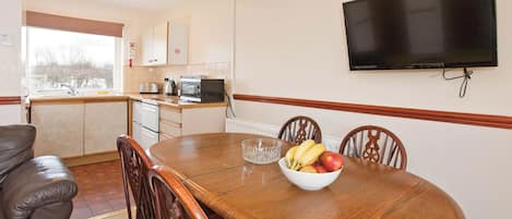 A dining area with a wooden table, chairs, a bowl of fruit, a television mounted on the wall, and a kitchen in the background.