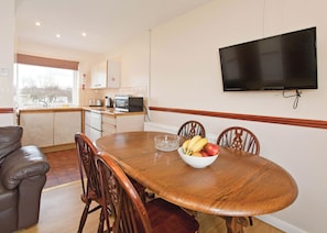 Un coin repas avec une table en bois, des chaises, un saladier rempli de fruits, un téléviseur fixé au mur et, au fond, une cuisine.