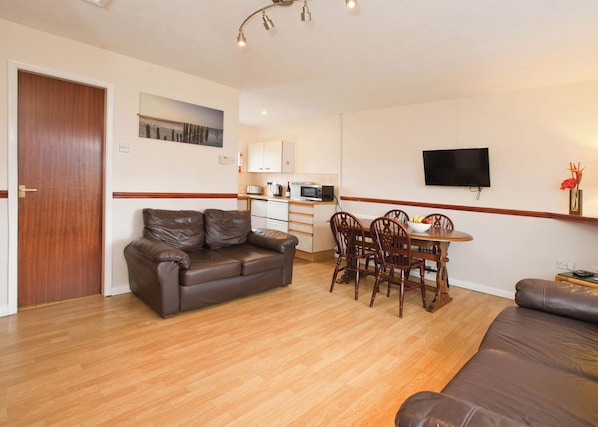 A living room with a sofa, a dining table with chairs, a kitchen area, and a television mounted on the wall. - Bungalow with one double and one twin. (Trearddur Bay, Holyhead)