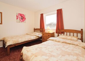 Een kamer met twee houten bedden, een nachtkastje met een lamp, een raam met gordijnen, en een schilderij aan de muur.
