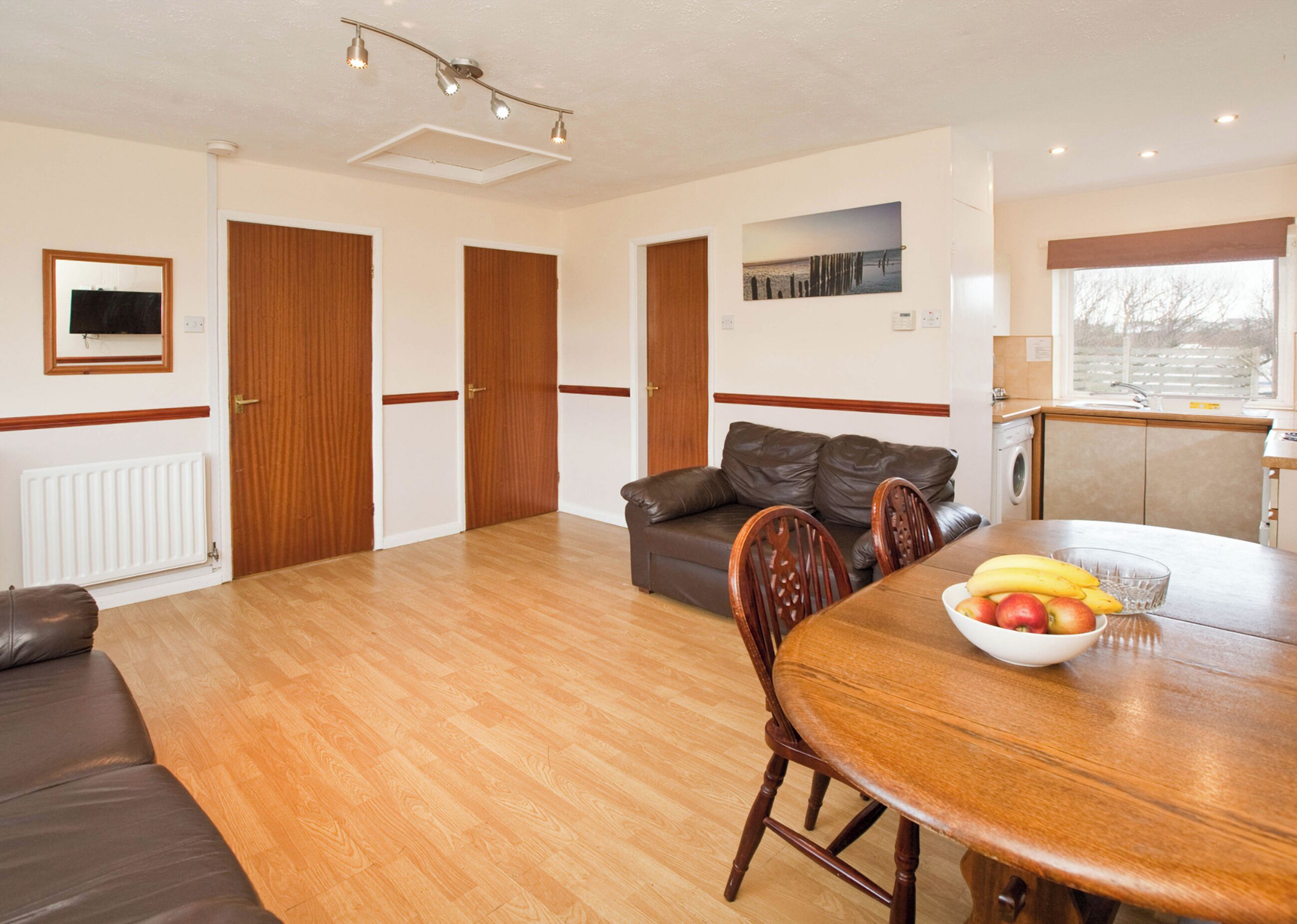 A spacious living area with a wooden floor, a brown leather sofa, a dining table with chairs, and a kitchen area with a window.