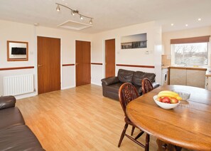 Een ruime woonkamer met houten vloer, een bruine leren bank, een eettafel met stoelen en een keuken met raam.