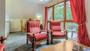 A room with two wooden armchairs, a wooden table, a window with red curtains, and a view of greenery outside. - Highland Ranch Lodge (Delny, Nr Invergordon)