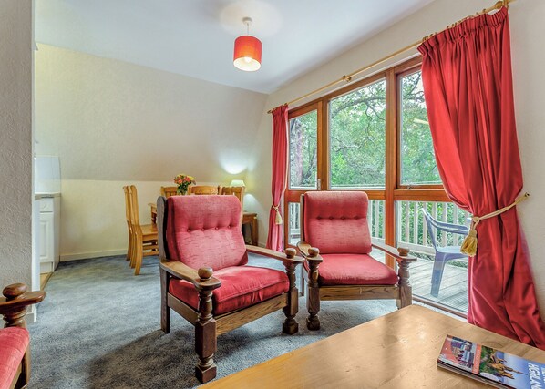 A room with two wooden armchairs, a wooden table, a window with red curtains, and a view of greenery outside. - Highland Ranch Lodge (Delny, Nr Invergordon)