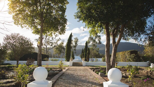 Un sendero en un jardín conduce a una estructura blanca con esculturas esféricas en los extremos, rodeado de árboles y vistas de colinas distantes.