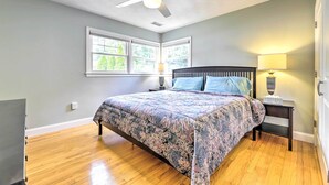 Un dormitorio con cama, ventilador de techo, mesita de noche con lámpara y ventanas con vistas a árboles.