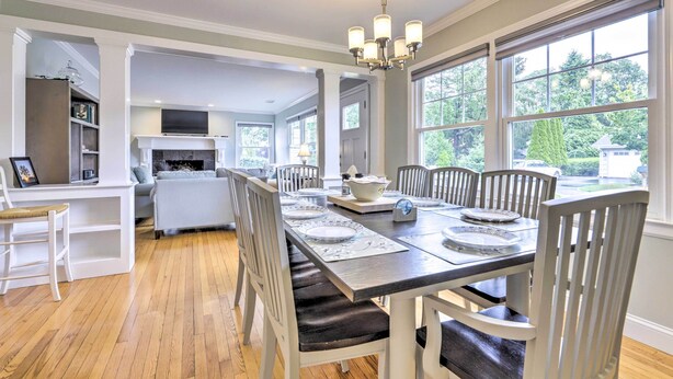 Comedor con una mesa larga dispuesta para seis personas, rodeada de sillas blancas, un candelabro y ventanas grandes con vistas a follaje.