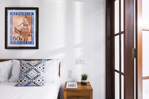 Cama bem arrumada com almofadas estampadas, uma pequena mesa de cabeceira de madeira com uma planta e um selo emoldurado da Indonésia.