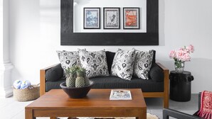 A living room with a black-framed wall art, patterned pillows, a wooden coffee table with a pineapple bowl, and a vase with pink flowers. - Villa Anahera (Seminyak)