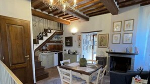 Un interior rústico con vigas de madera a la vista, un candelabro, una mesa de comedor con sillas, una chimenea y una escalera que lleva a una cocina.