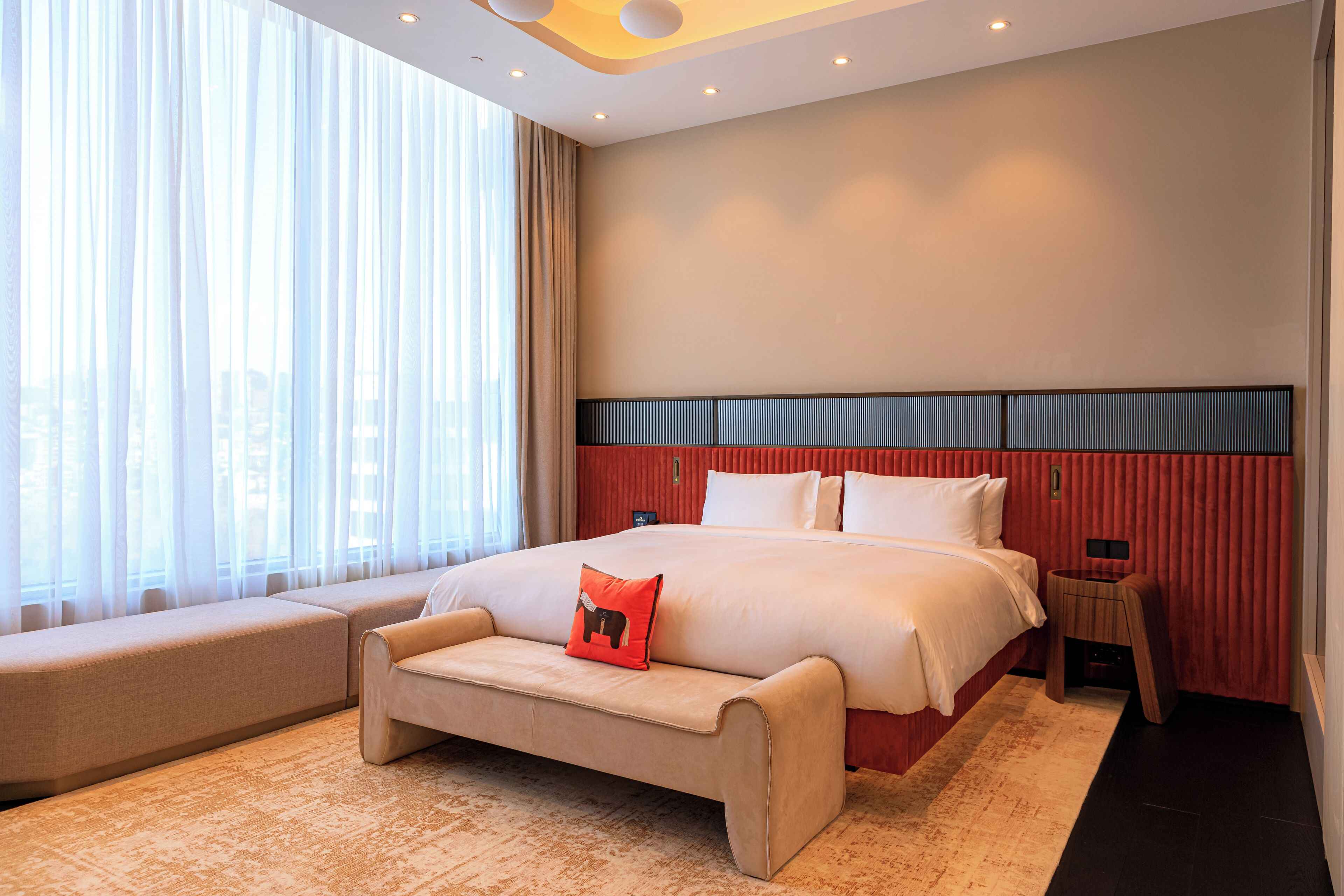 A modern hotel room with a large bed, a bench, a nightstand, and a view of the city through sheer curtains.