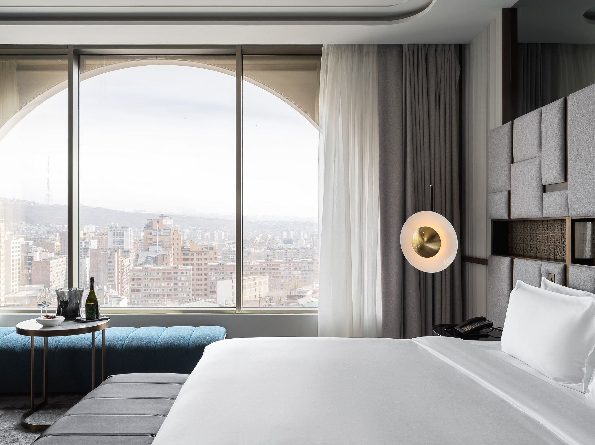 A modern hotel room with a large bed, a seating area with a small table, and a city view through large windows.
