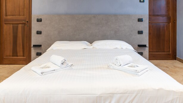 A neatly made bed with a grey headboard, two white towels, and a wooden door on the left.