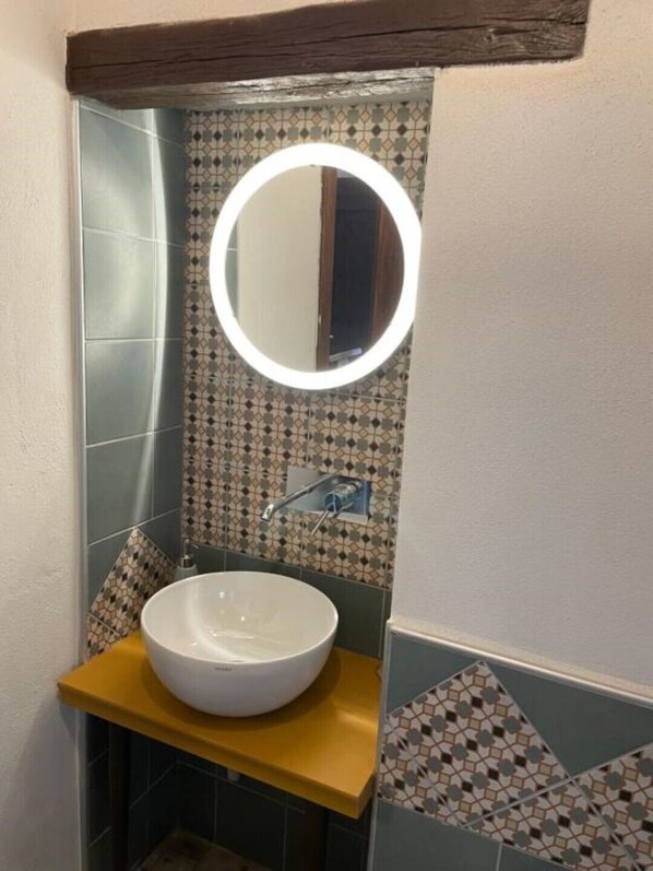 A bathroom with a white vessel sink, a round mirror with a light, and patterned tiles. - The Bastion Attic (Pitigliano)