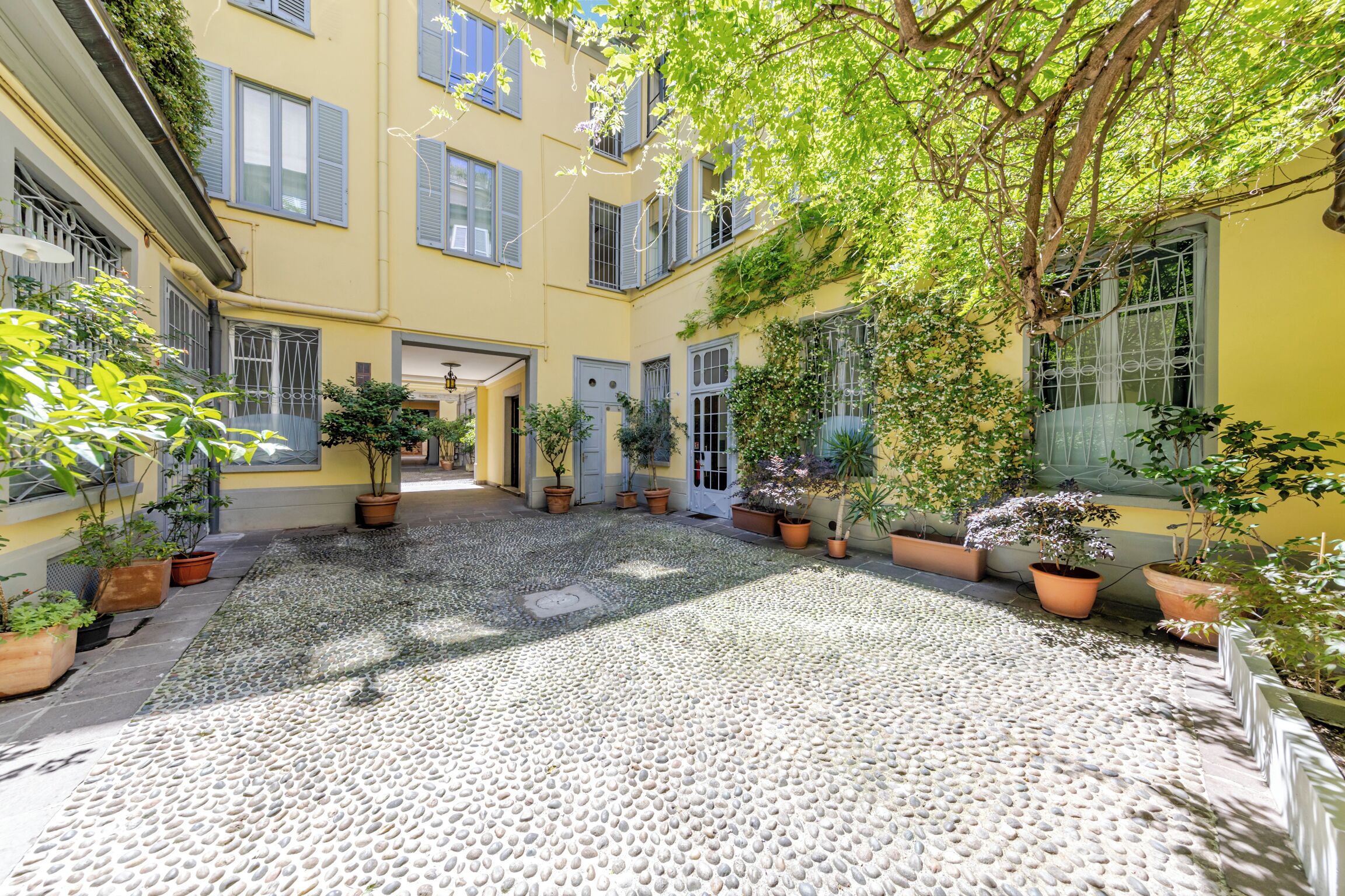 A courtyard with a cobblestone floor, potted plants, and a yellow building with multiple windows and doors.