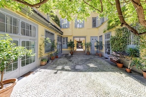Un patio con suelo de adoquines, plantas en macetas y un edificio con ventanas y contraventanas grandes.