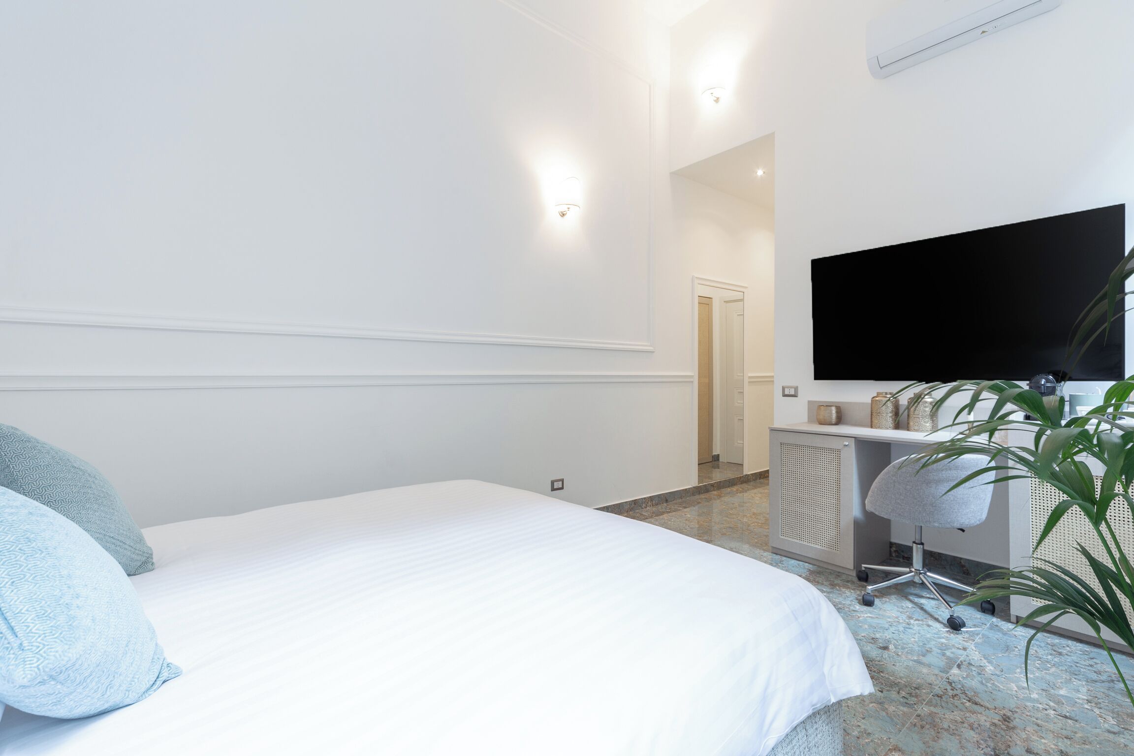 A hotel room with a bed, a desk with a chair, a television, and a plant.