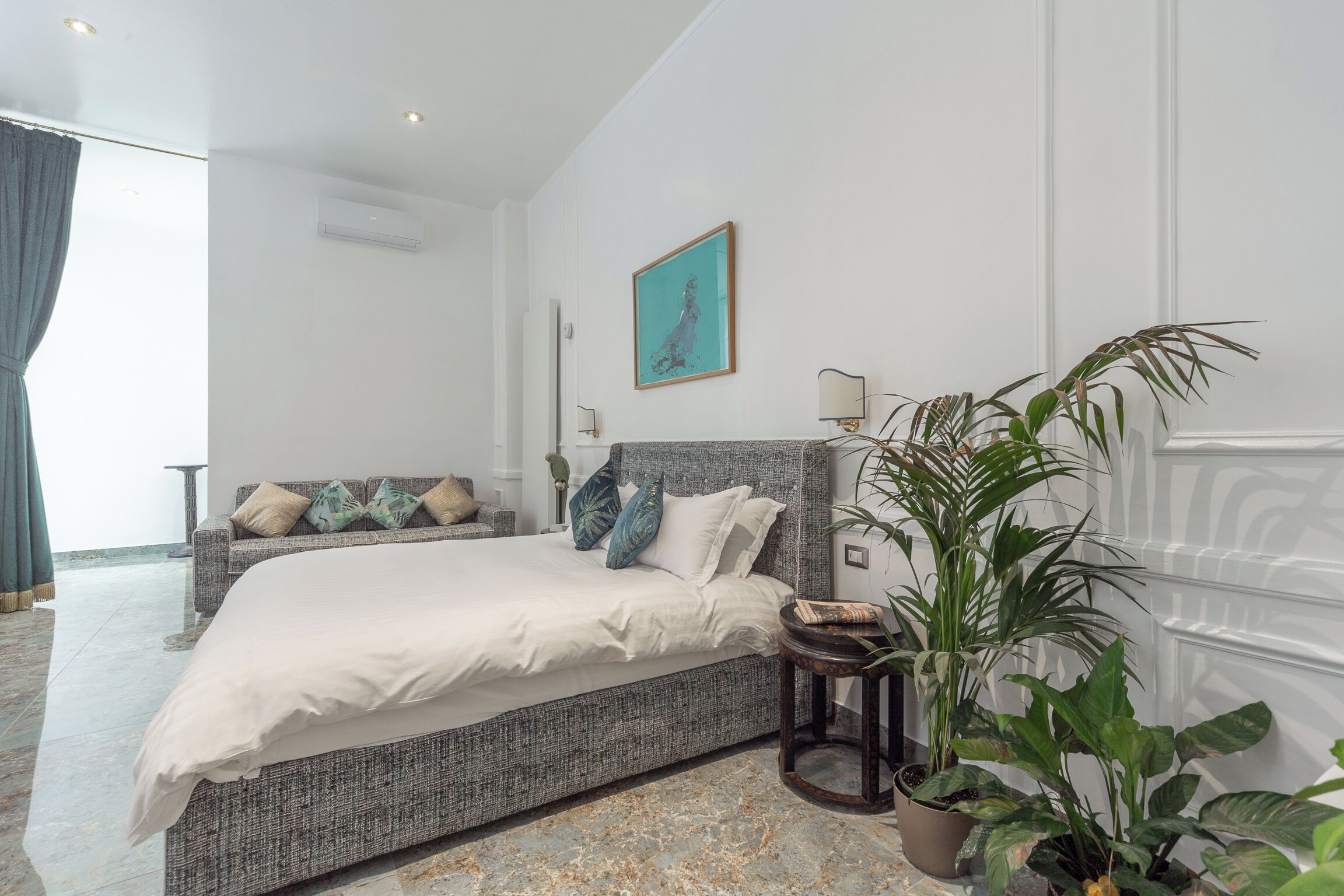 Un dormitorio con una cama grande, un cabecero gris, una planta en maceta y un cuadro en la pared.