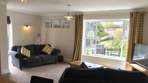 A living room with a sofa, a dining table with fruit, a TV, and a window with curtains. - Lovely 3-bed House, Burnham Market, North Norfolk (King's Lynn)