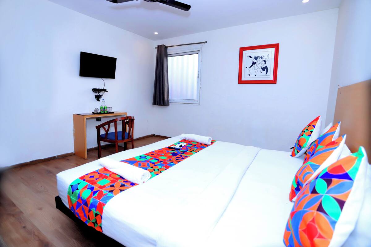 Habitación de hotel con cama, escritorio, silla, televisor, ventana con cortinas e imagen enmarcada en la pared.