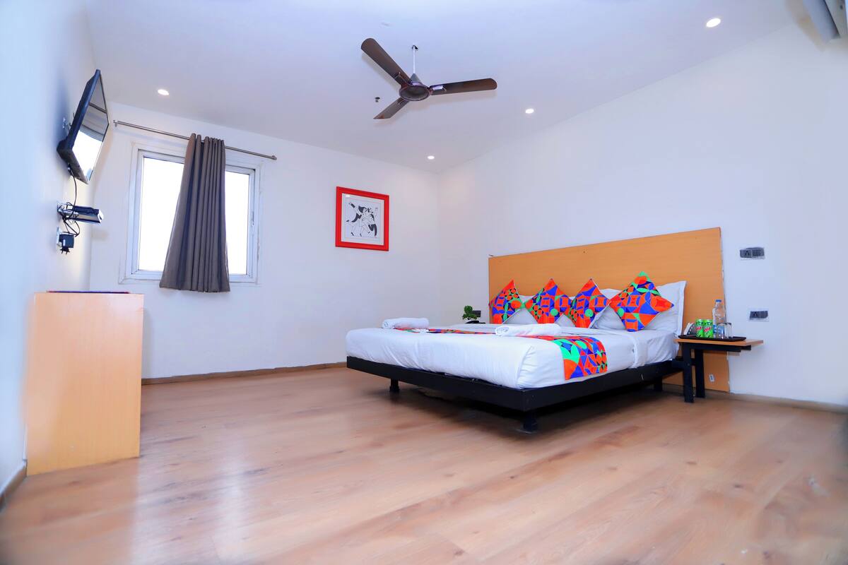 Un dormitorio con cama, ventilador de techo, ventana con cortinas, mesita de noche con una botella y televisor instalado en la pared.