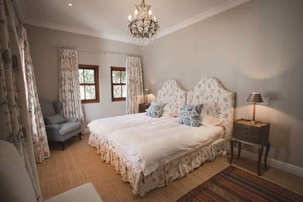 A bedroom with a large bed, a chair, a nightstand, a chandelier, and floral curtains.