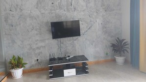 A living room with a flat-screen TV mounted on a marble wall, a black entertainment unit with electronic devices, and two potted plants. - Charming House in Matugga, Kampala, Uganda (Matugga)