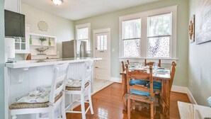 A kitchen with white cabinetry, a dining area with wooden chairs and a table, and a large window with blinds. - Coastal Blue Cottage is Steps to Beach and Seawall (Galveston)
