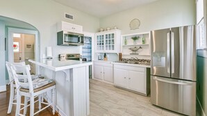 A modern kitchen with white cabinets, stainless steel appliances, and a central island with bar stools. - Coastal Blue Cottage is Steps to Beach and Seawall (Galveston)