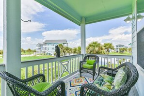 A covered porch with wicker chairs and a colorful rug, overlooking a grassy area with a building in the background.
