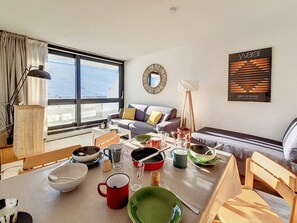 A modern living room with a dining table set for four, a sofa, a coffee table, and a large window offering a view of the outdoors.
