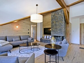 Una sala de estar moderna con techo alto, vigas de madera a la vista, chimenea de piedra y un conjunto de sofá gris.