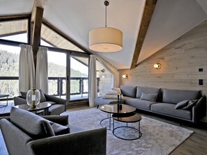 Una sala de estar moderna con un sofá, sillones, una mesa de centro y un amplio ventanal que ofrece vistas a una zona boscosa.
