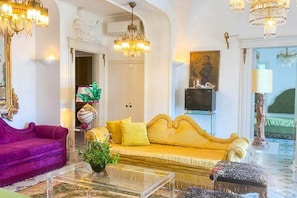 Una habitación con un sofá morado, un sofá amarillo, una mesa de centro de vidrio y un candelabro.