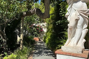 Un sendero en un jardín con una estatua a la derecha, rodeado de follaje y plantas en flor.