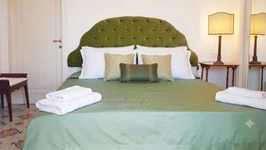 Ein ordentlich bezogenes Bett mit einem grünen Kopfteil, weißen Kissen und einer grünen Tagesdecke. Neben dem Bett steht ein Holzhocker und ein kleiner Tisch mit einer Lampe.