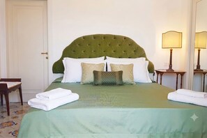 A neatly made bed with a green headboard, white pillows, and a green bedspread. There is a wooden chair and a small table with a lamp beside the bed.