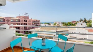 Un balcon avec des chaises turquoise et une table, donnant sur une zone côtière avec des bâtiments et un ciel dégagé.