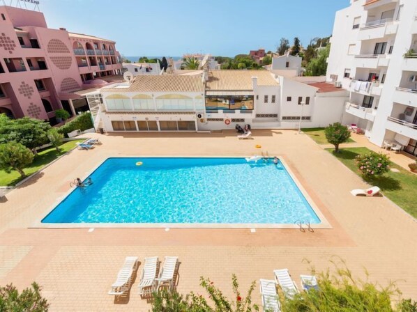 Una piscina rectangular con sillones de descanso y una zona cubierta, rodeada de edificios con balcones y una vista despejada al mar.