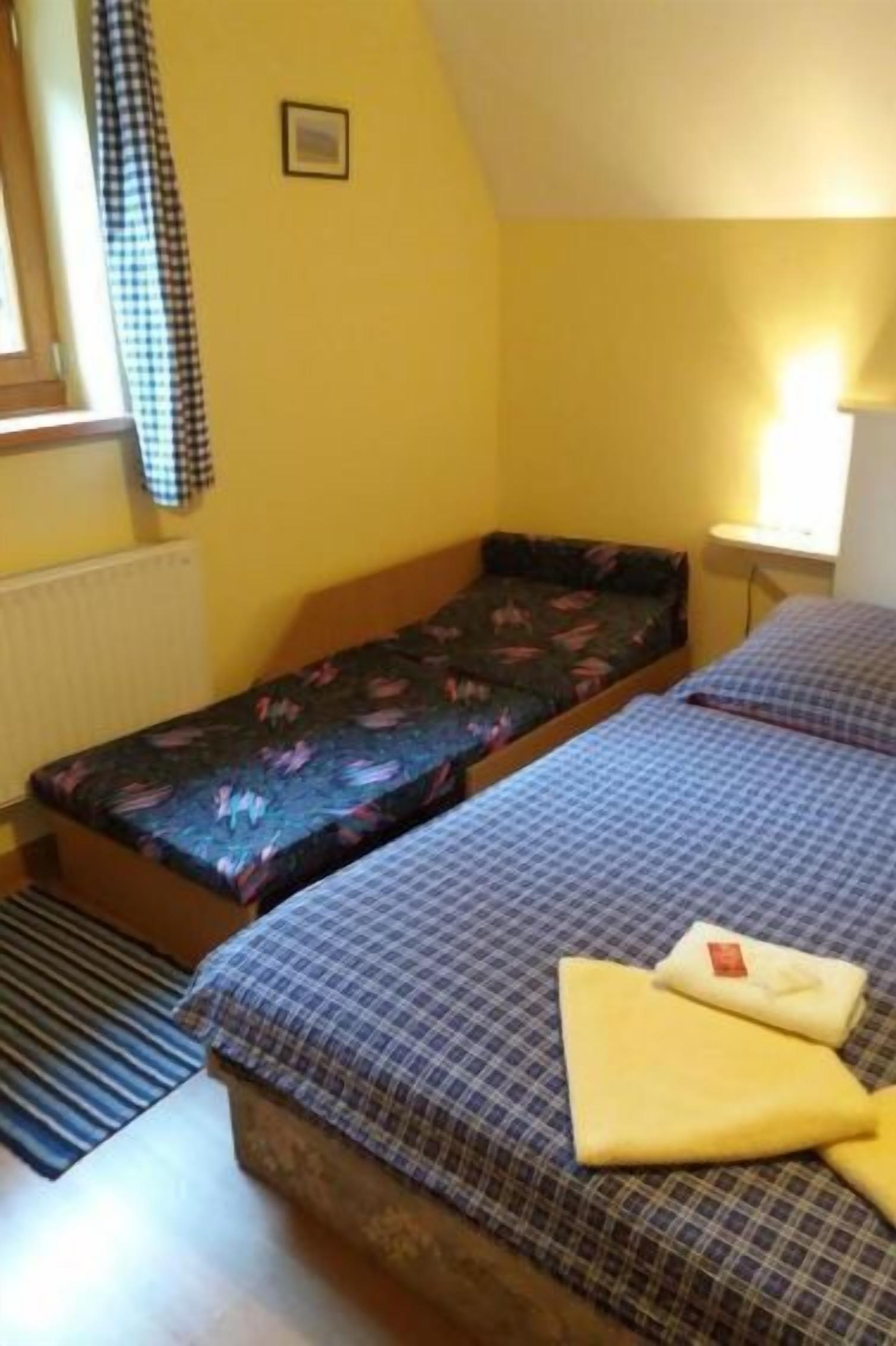 A small, single-room accommodation with two beds, a radiator, a window with a checkered curtain, and a bedside table with a lamp.