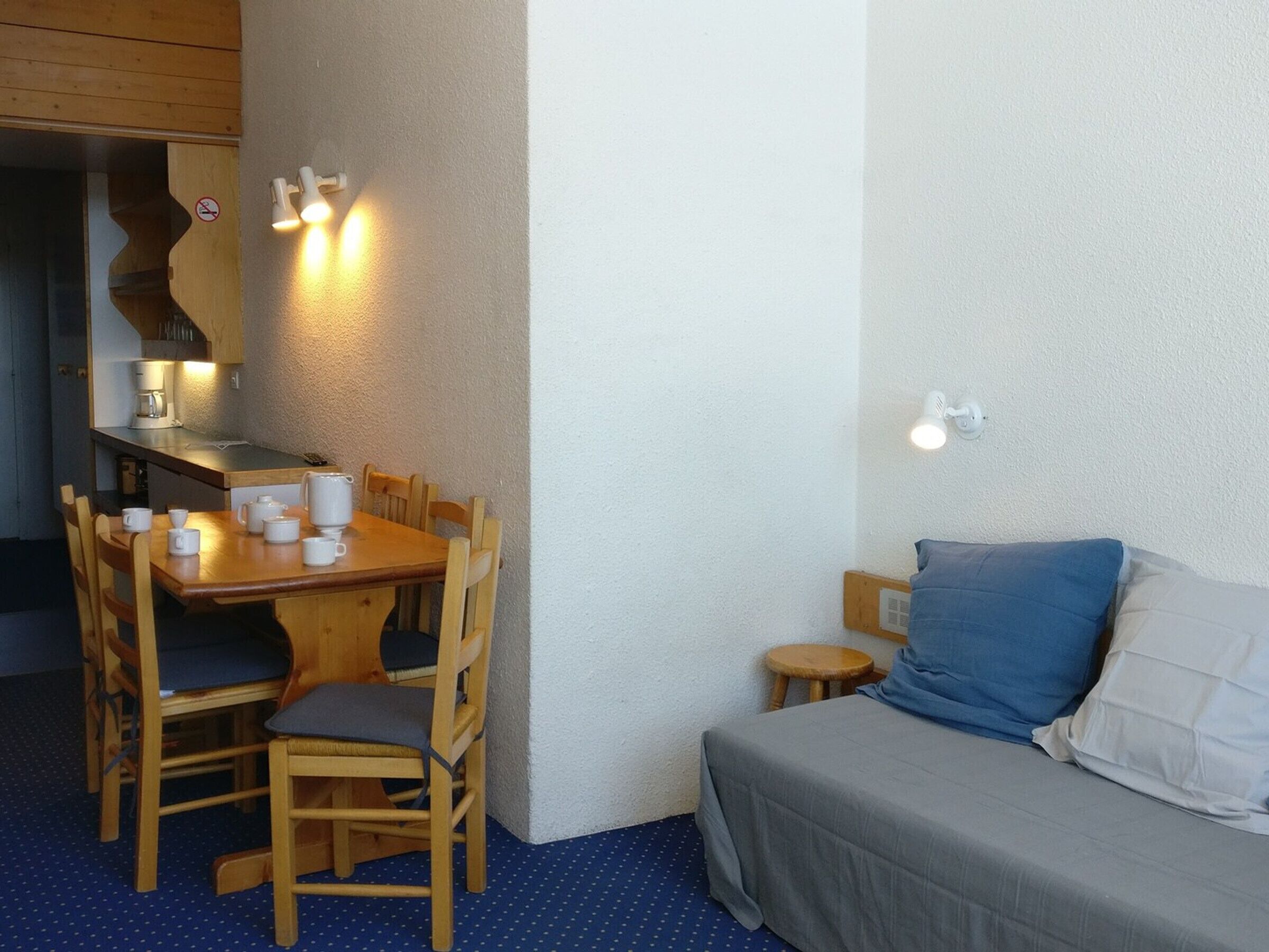 Ein kleiner, gemütlicher Raum mit einem Holzespeisetisch und Stühlen, einem kleinen Hocker, einem Bett mit grauer Decke und einer Wandleuchte.